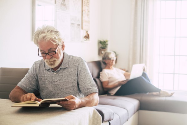 Senior man reading book as his wife uses laptop in living room