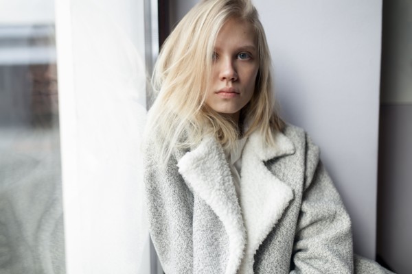 young woman in wool coat leaning against wall by window