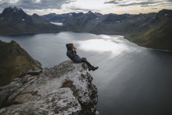 Norway, Senja, Man taking photo sitting on edge of steep cliff on top of mountain Segla
