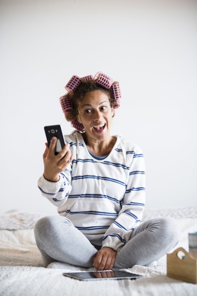 Woman with hair curlers sitting on bed and taking selfie