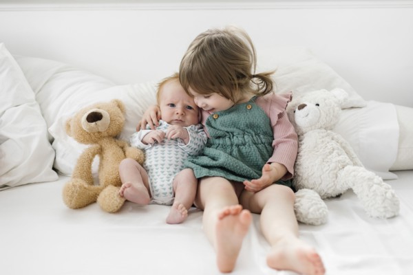 Siblings (2-5 months, 2-3) sitting on bed with teddy bears