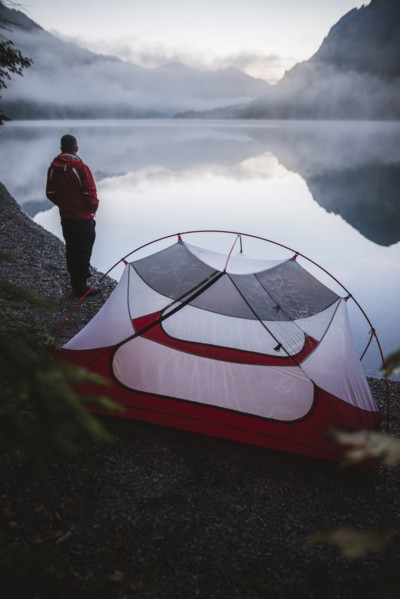 Austria, Plansee, Young man standing near tent and looking at Plansee lake at sunrise