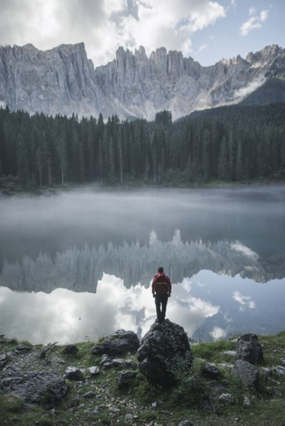 Italy, Carezza, Young man standing on rock at Lago di Carezza in Dolomite Alps at dawn