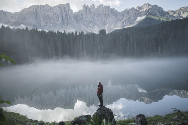 Italy, Carezza, Young man standing on rock at Lago di Carezza in Dolomite Alps at dawn