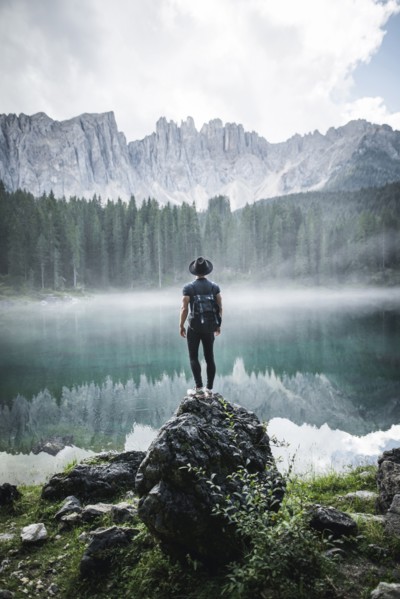 Italy, Carezza, Young man standing on rock at Lago di Carezza in Dolomite Alps at dawn