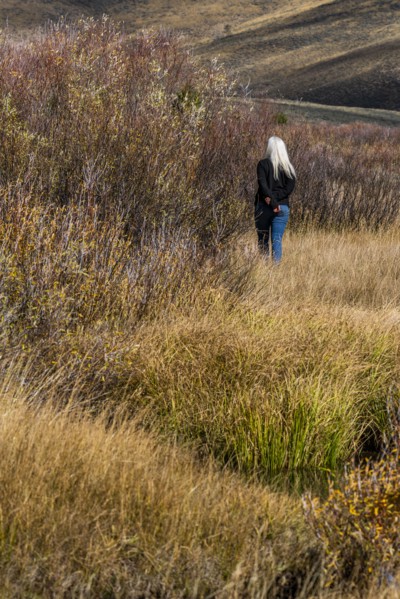 Senior woman with long white hair walking by stream among grass in non urban landscape