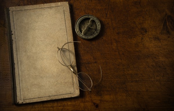 Old book with blank cover resting on old wooden desk top with old eyeglasses and small sundial