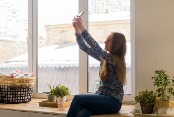 Caucasian woman sitting at window posing for cell phone selfie