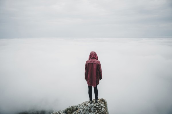 Distant Caucasian woman standing on rock watching fog on ocean