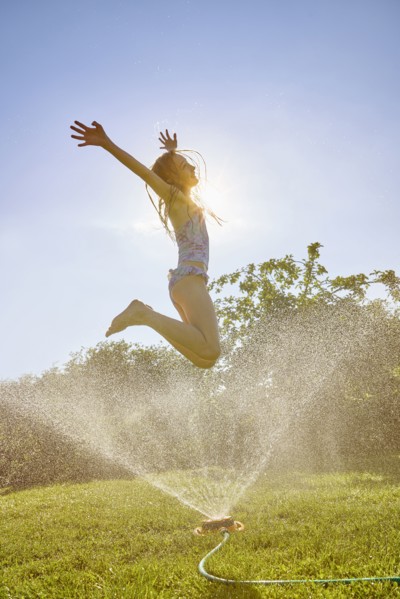 Caucasian girl jumping over backyard sprinkler