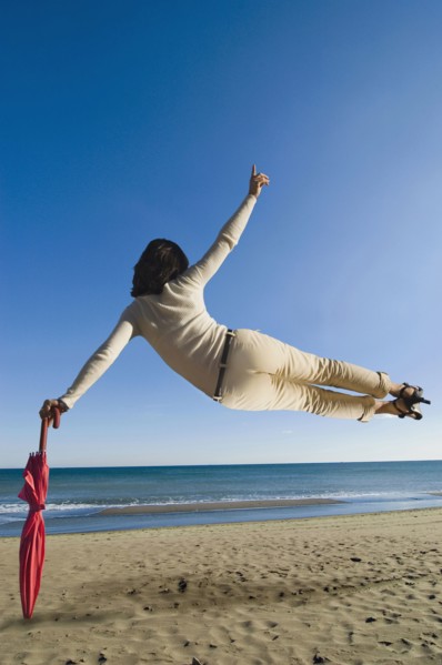 Woman jumping mid air on beach leaning on umbrella rear view
