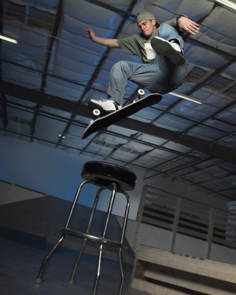 Caucasian man jumping on skateboard over stool indoors