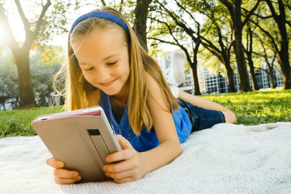 Smiling Caucasian girl laying on blanket in park reading digital tablet