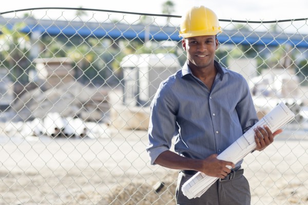 Black architect in hard hat holding blueprints near fence