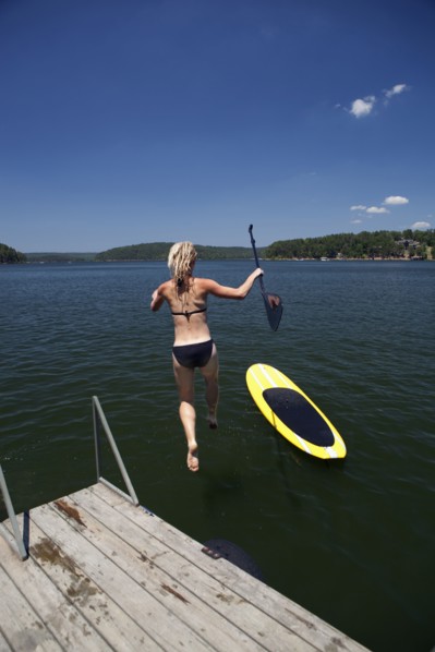 Caucasian woman jumping in lake next to paddleboard