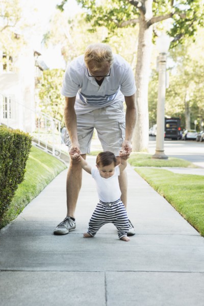 Father helping baby son walk on sidewalk