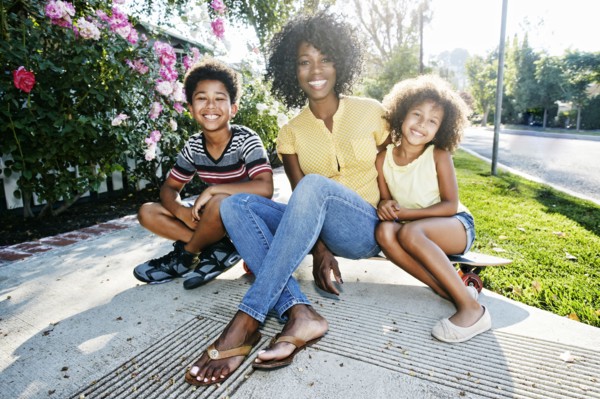 Smiling mother and children sitting on skateboard on sidewalk