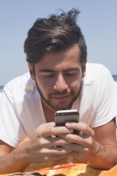 Hispanic man laying on blanket at beach texting on cell phone