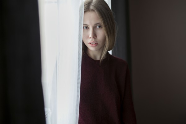 Serious Caucasian woman standing near curtain at window