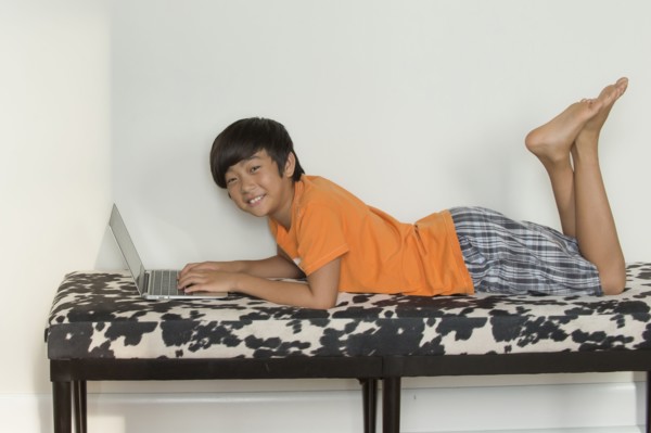 Smiling Asian boy laying on bench using laptop