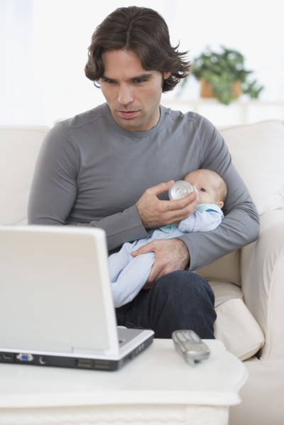 Hispanic father feeding baby and looking at laptop