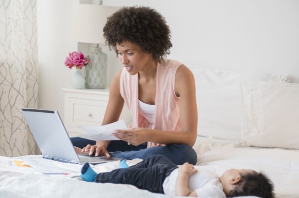 Mother paying bills on laptop with baby son on bed