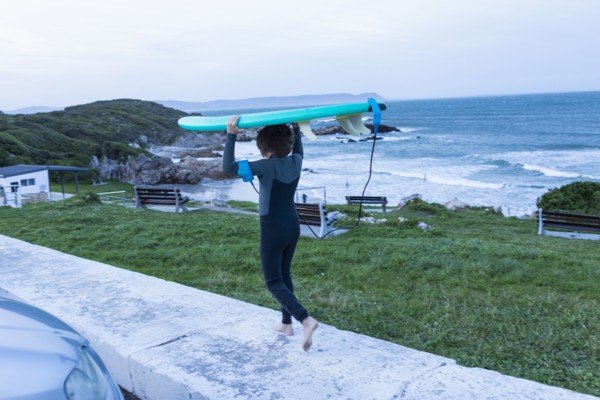 Boy carrying surfboard on head while walking on sidewalk at dusk in Voelklip Beach