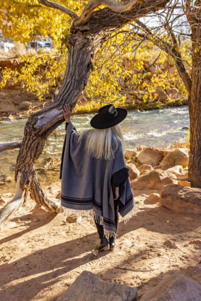 USA, Utah, Zion National Park, Rear view of woman in poncho and hat standing by tree along Virgin