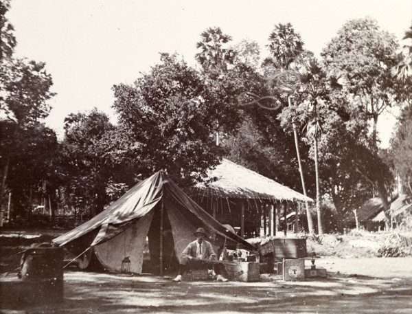 Camping in a village. A European man sits on an upturned crate outside his tent, brandishing a