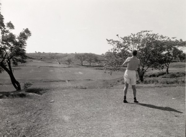 Playing golf, Rangoon. A lone golfer follows his shot, club still swung high in the air, on the