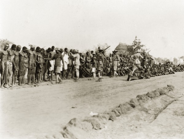 Sleeping sickness inspection. Sudanese men and boys stand and sit in a line on a roadside awaiting