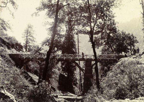 Bridge on the way to Kashmir. A distant figure peers over the side of a wooden road bridge,