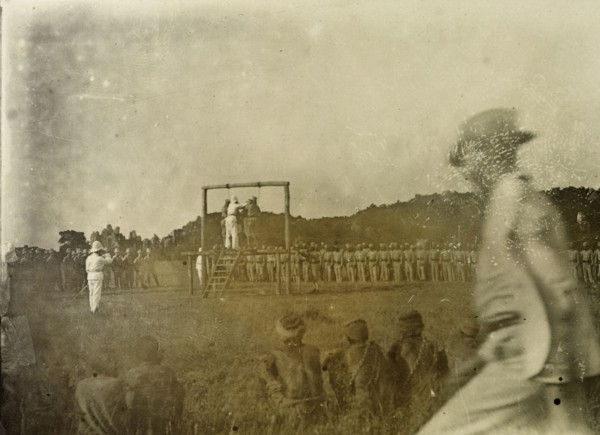 Colonial execution. An executioner prepares for two African men to be hung from a wooden scaffold,