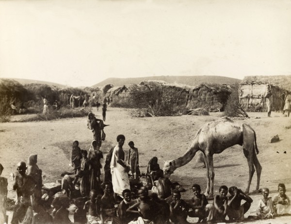 Village scene at Aden. A cluster of people, many of them children, stand or squat on the sand in