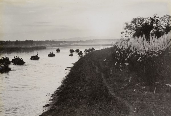 Return to camp. Mounted on elephants, King George V's royal hunting party crosses a river at sunset