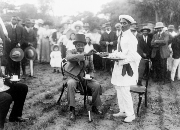 Tea and cake at a royal reception. Dressed in a Western-style suit and top hat, an African chief