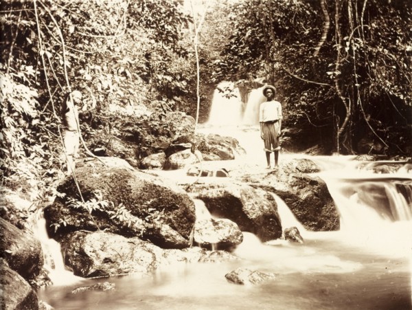 Jungle river in Bioko. Two young men stand on a rock formation straddling a jungle river. The slow