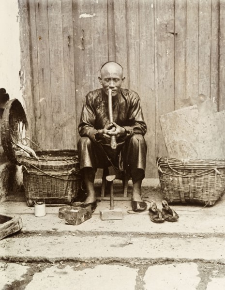 An itinerant shoemaker, Hong Kong. An itinerant shoemaker sits on a stool, smoking a long pipe at