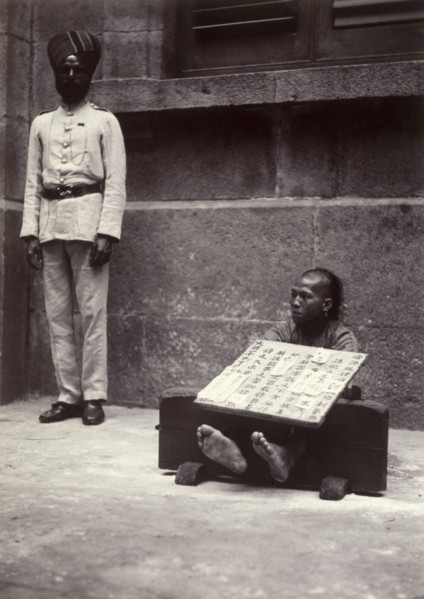 Petty criminal in the stocks, Hong Kong. A petty criminal sits with his ankles restrained in
