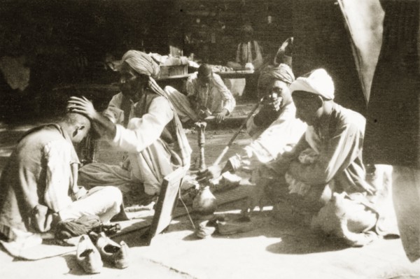 Barber's stall on a street in Peshawar. A man sits cross-legged as he has his head shaved at a