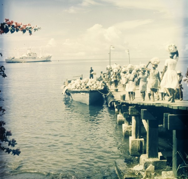 Maritime trade, Dominica. A group of women walk along a wooden pier carrying sacks on their heads,