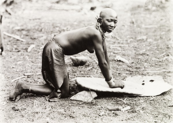 Grinding millet. A Kikuyu woman, naked from the waist up, kneels on the ground as she grinds millet