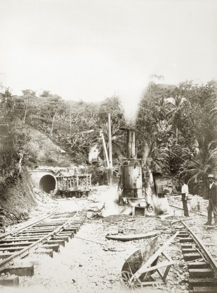 Railway construction site. Labourers stand on the tracks beside steam-operated machinery at a