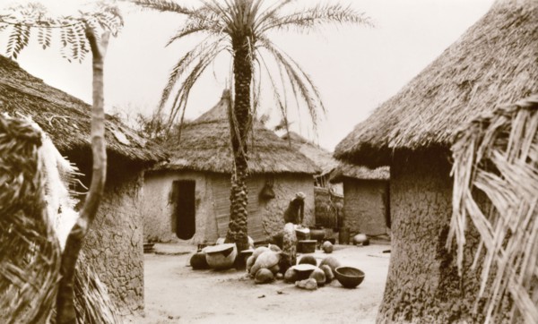 Interior of a Nigerian village. A woman undertakes domestic chores outdoors in the centre of a