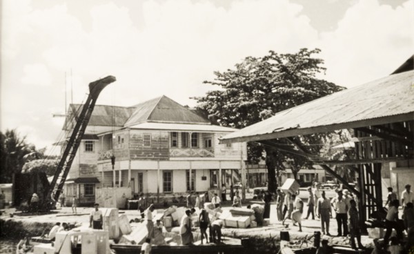Unloading goods at Apia wharf. Crates of goods are unloaded from barges at a wharf in the port city