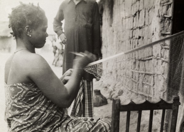 Repairing fishing nets. A Ghanaian woman in traditional dress sits outside a mud-walled building as
