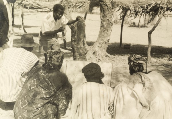 Adult literacy campaign, Gold Coast. A student teacher helps men to read from a board during a