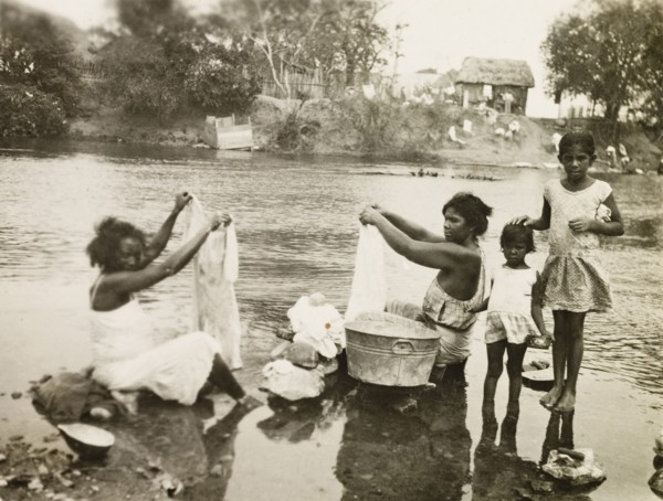 Washing clothes in a stream. Two female plantation workers, accompanied by two young girls, sit in