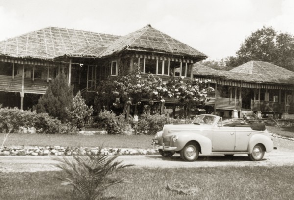 A stilted bungalow in British Malaya. A small car is parked in the driveway outside Dr Tweedie's