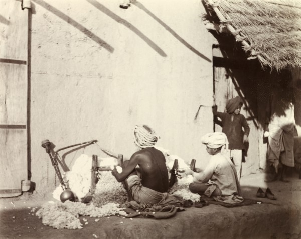 Indian cotton weavers. Two Indian men sit weaving cotton on the ground outside a mud-walled